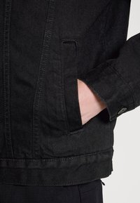 Czarna kurtka jeansowa z kołnierzem, zapięciem na guziki i przednimi kieszeniami. Charakteryzuje się fakturowaną tkaniną oraz kontrastującym przeszyciem na dole i mankietach.