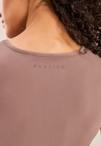 Top activewear in tessuto malva tenue con texture liscia, caratterizzato da uno scollo arrotondato e logo in rilievo vicino alla spalla.