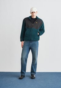 Fleecepaita, jossa on petroolinvärinen runko ja mustat olkapäät, oranssi vetoketjusomistus, yhdistetty sinisiin farkkuihin ja mustiin kenkiin, päässä valkoinen lippalakki.