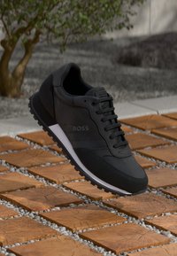 Zwarte sport sneaker met een glad materiaal, gestructureerde accenten, logo aan de zijkant, vetersluiting en een witte tussenzool.
