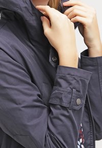 Navy blue water-resistant jacket featuring a high collar, zippered front, and buttoned sleeve cuffs. Smooth fabric with minimalistic design.