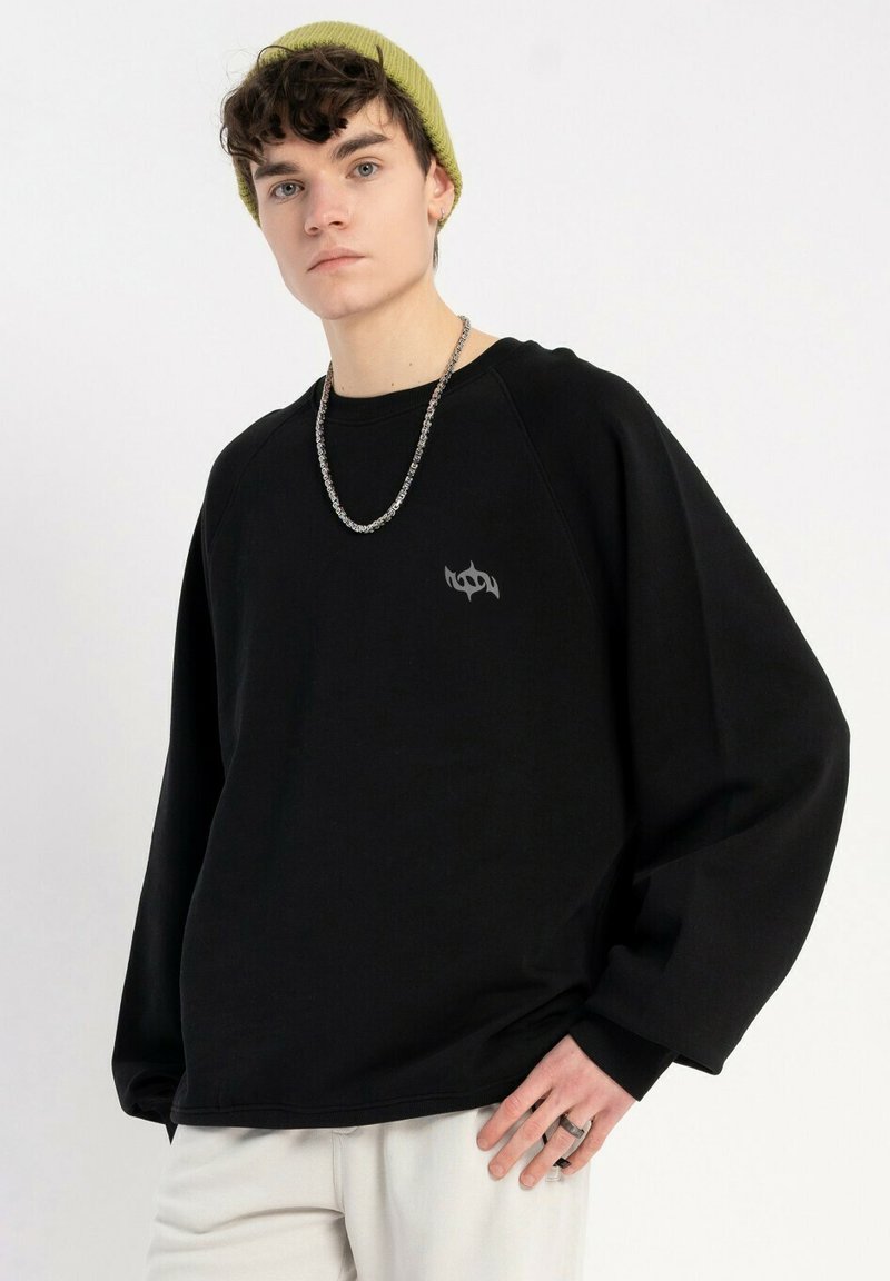 Czarny oversize sweatshirt z subtelnym logo na piersi, wykonany z bawełny; noszony z zieloną czapką beanie i srebrnym naszyjnikiem z łańcuchem.