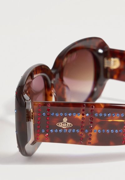 Lunettes de soleil à monture écaille avec des verres arrondis et des branches décorées de strass rouges et bleus ainsi qu’un petit emblème doré.