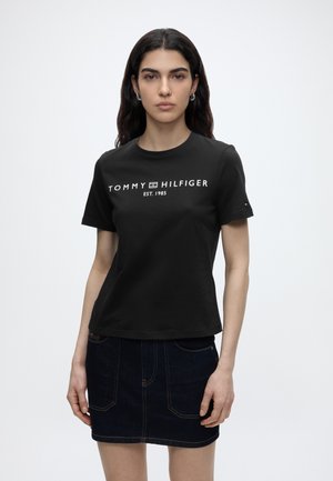 Νέα γυναίκα με μακριά σκούρα μαλλιά που φοράει μαύρο t-shirt Tommy Hilfiger και σκουρόχρωμη τζιν φούστα σε μονόχρωμο ανοιχτό φόντο.