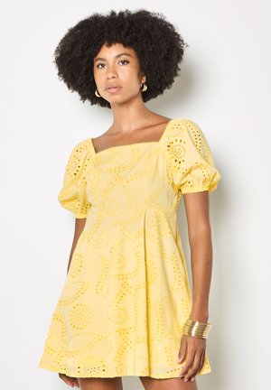 Frau mit lockigem Haar trägt ein gelbes Kurzarmkleid mit quadratischem Ausschnitt aus Lochstickerei sowie goldene Creolen und ein Armband.