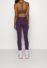 Hnědá sportovní podprsenka s designem na zádech, spárovaná s fialovými legínami s vysokým pasem. Na nohou jsou bílé sportovní boty s oranžovými akcenty.
