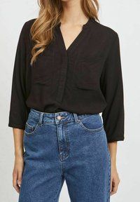 Blusa nera a maniche lunghe con scollo a V e due tasche sul petto, abbinata a jeans in denim blu a vita alta con chiusura a bottone.