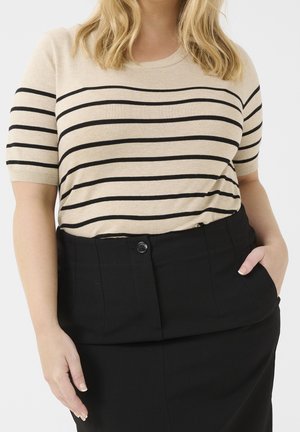 Frau trägt beige Kurzarmoberteil mit schwarzen horizontalen Streifen, in einen hoch taillierten schwarzen Rock mit Knopf und Taschen gesteckt.