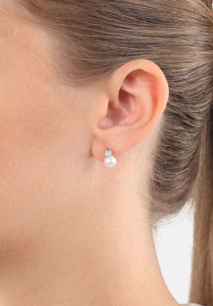 Oreja de mujer con cabello castaño claro recogido, usando pendientes de plata en forma de botón con una sola perla y una pequeña gema clara encima.