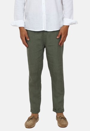 Uomo che indossa pantaloni verde oliva con risvolti, una camicia bianca a maniche lunghe abbottonata e scarpe stile mocassino color cammello, in piedi con le mani rilassate.