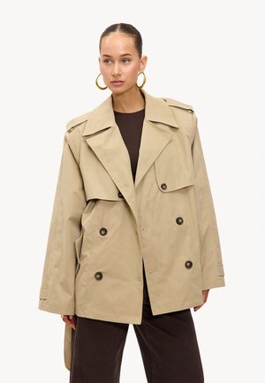 Femme portant un trench-coat beige à double boutonnage, un haut marron foncé et un pantalon, avec des boucles d'oreilles en or, se tenant devant un fond blanc uni.