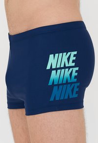 Marina blå badbyxor gjorda av stretchmaterial, med en staplad gradient Nike-logga i ljusblått på sidan. Sitter åtsittande.