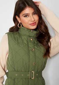 Zelená prošívaná vesta s vysokým límcem, zlatými doplňky a opaskem v pase. V kombinaci s svetrem z světle barvené příze.