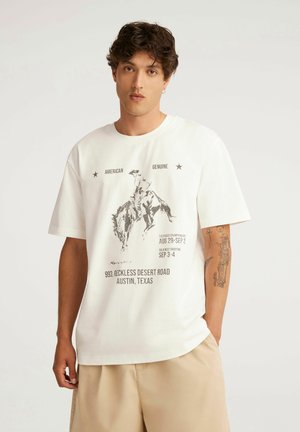 Młody mężczyzna z krótkimi kręconymi włosami w białym T-shircie z nadrukiem rodeo i beżowych szortach, stoi na jednolitym tle.