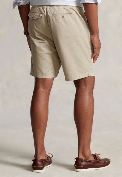 Medvilniniai khakio spalvos šortai tiesiu kirpimu, su dviem užpakalinėmis kišenėmis su sagomis ir lygiu paviršiumi, derinami su rudos spalvos batais.