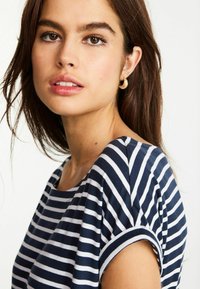 Marinblå och vit randig t-shirt med avslappnad passform och uppvikta korta ärmar. Modellen bär guldörhängen. Slät tygtextur synlig.