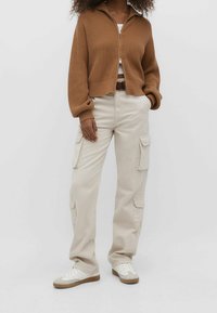 Bruine geribbelde ritstrui, beige cargo broeken met zijzakken en witte sneakers met bruine rubberzolen. Casual, relaxte pasvorm.