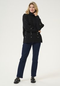 Svart stickad tröja med hög hals, prydd med pärlaccenter och en strukturerad design. Matchad med mörkblå denimbyxor och svarta skor.