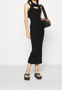 Mustat ilman hihoja midi-mekko, jossa on leikkaus yksityiskohtana rinnassa, istuva siluetti ja ribattu pinta. Yhdistettynä alustakevyisiin flip-flop-kenkiin.