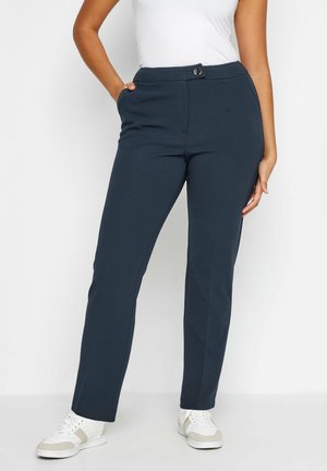 Femme portant un pantalon bleu foncé sur mesure avec des poches latérales, un débardeur blanc et des baskets blanches, debout devant un fond uni.