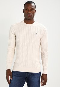 Suéter de punto cableado en color blanco roto, con mangas largas, cuello redondo, puños y dobladillo acanalados, que presenta un pequeño logo oscuro en el pecho.