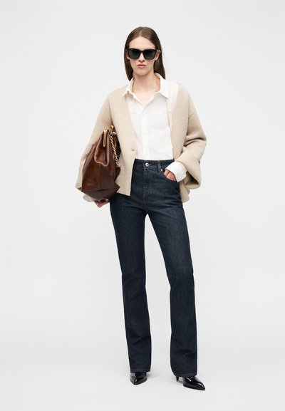 Woman wearing beige cardigan, white shirt, dark jeans, black pointed boots, holding large brown handbag with gold chain, and wearing black sunglasses.