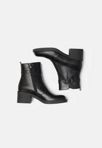 Bottines en cuir noir avec un bout arrondi, un talon block, une fermeture éclair sur le côté et un détail de sangle avec une boucle. Texture lisse, design minimaliste.