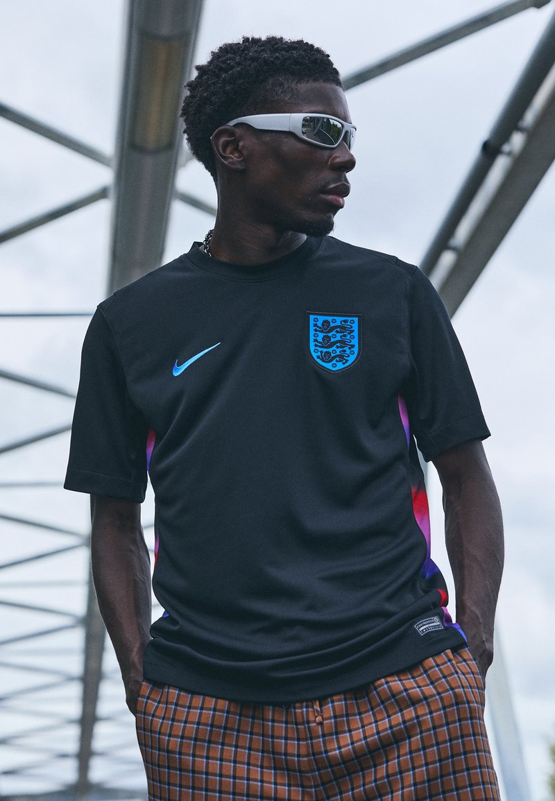 Crna Nike majica s plavim grbom i logotipom, kratki rukavi i gradijentni ljubičasti akcenti. Smešane karirane hlače s plavim linijama.