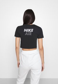 Kvinna med långt mörkt hår som bär en svart croppad t-shirt med texten "NIKE AIR" på ryggen och vita höga byxor, vänd bort.