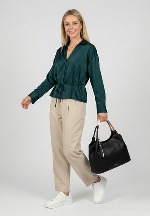 Usmívající se žena v tmavě zelené blůze a béžových kalhotách, držící velkou černou kabelku, stojící před jednobarevným světlejším pozadím.