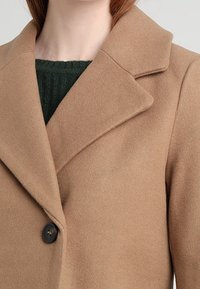 Brun ylleblandad kappa med en snedskuren krage, en svart knapp och en mjuk textur, lagd över en mörkgrön ribbad stickad topp.