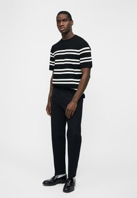 Άνδρας που στέκεται, φοράει μαύρο και λευκό ριγέ κοντομάνικο πουκάμισο, μαύρο παντελόνι, λευκές κάλτσες και μαύρα μοκασίνια σε απλό λευκό φόντο.