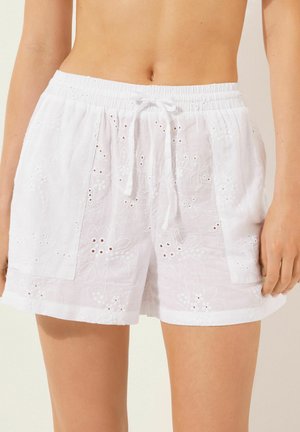 Weiße Shorts mit floraler Stickerei, Lochmuster-Details, einem elastischen Bund mit Kordelzug und Seitentaschen. Der Leinwandstoff ist sichtbar.