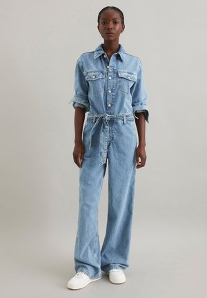 Une combinaison en denim bleu clair avec un col, des manches longues, des poches poitrine et une ceinture à nouer, présentant des jambes larges et une coupe décontractée.