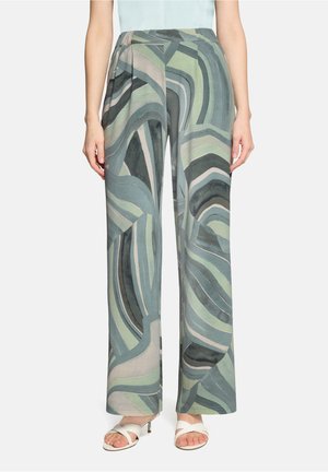 Femme portant un pantalon à jambes larges avec un motif abstrait ondulé vert, bleu et gris, accompagné de sandales blanches à talons.