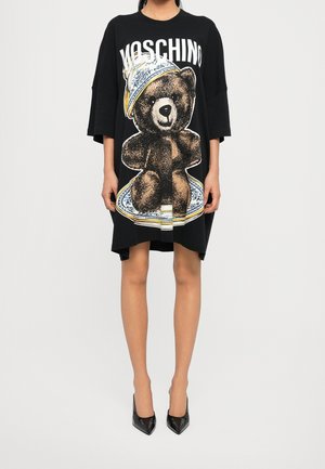 Kobieta ubrana w czarną, oversize sukienkę Moschino z dużą ilustracją misia pluszowego i czarne buty na wysokim obcasie.
