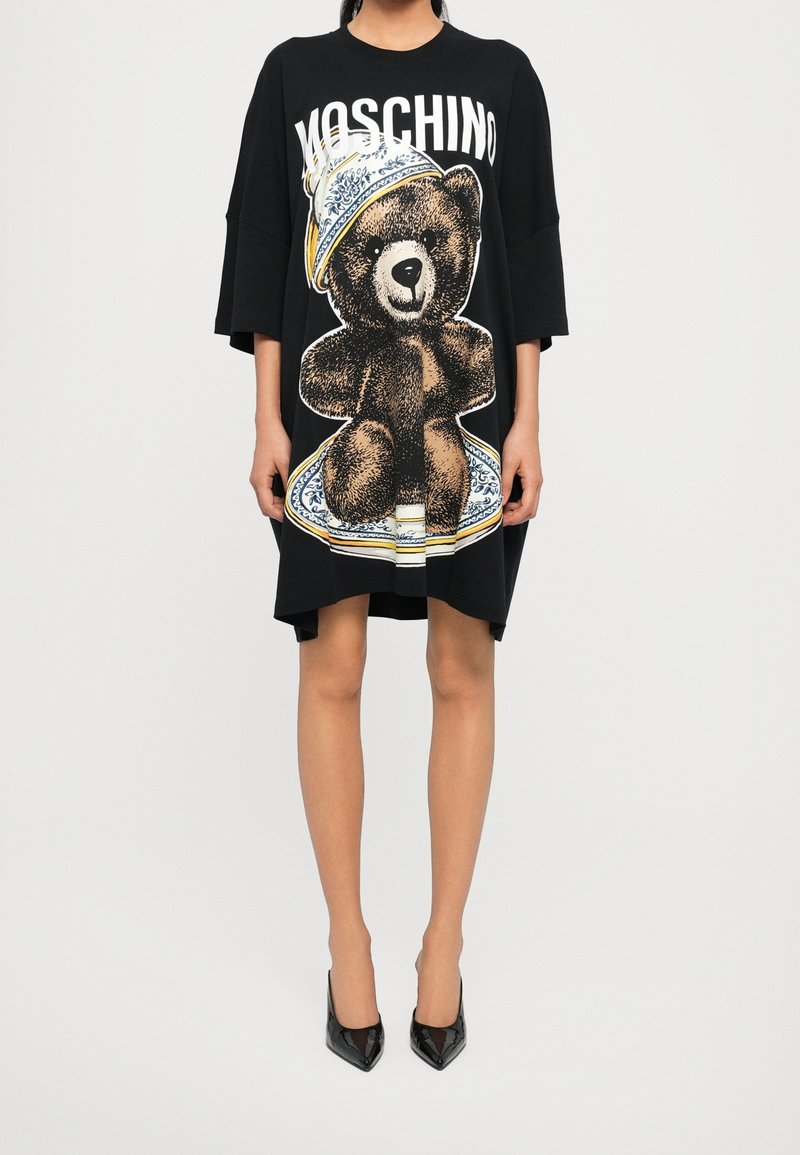 Femme portant une robe noire Moschino oversize avec une grande illustration d'ours en peluche et des chaussures à talons noires.