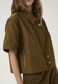 Braune Kurzarmjacke aus strukturiertem Stoff. Sie verfügt über einen Kragen, Vorderknöpfe mit Druckverschluss und eine seitliche Reißverschlusstasche.