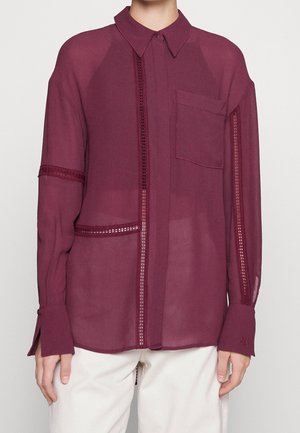 Osoba ubrana w przezroczystą bordową bluzkę z długim rękawem, zdobioną koronką, z kieszenią na piersi oraz białe spodnie na jednolitym tle.