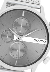 Montre métallique argentée avec trois sous-cadrans sur un cadran texturé gris. Elle est dotée d'un bracelet en maille et d'un logo affiché horizontalement.