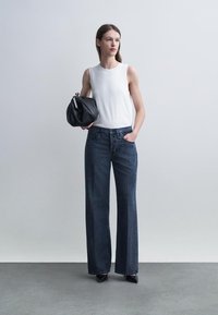 Femme portant un haut blanc sans manches et un jean large foncé tenant un sac à main noir, debout sur un sol gris devant un mur blanc.