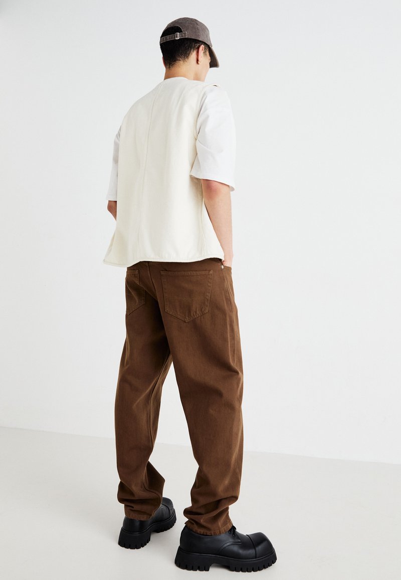 Man in een beige mouwloos vest, witte kort mouwen overhemd, bruine broek, zwarte stevige schoenen en een grijze pet, staand tegen een witte muur.