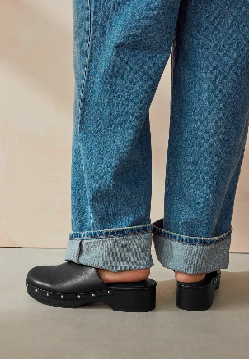 Personne portant un jean bleu retroussé et des sabots en cuir noir à enfiler avec des clous argentés, montrant les talons nus sur un fond beige.
