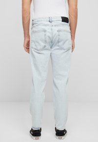 Svetlo modri denim jeans s ravnim krojem, petimi žepi in črnim etiketom na hrbtnem pasu; oblečeni z črnimi čevlji.