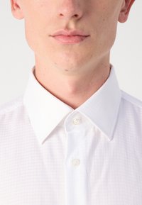 Chemise blanche à motifs avec un col pointu et un tissu lisse et texturé. Deux boutons visibles au niveau du col.