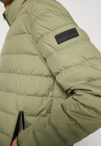 Giacca a vento verde con design trapuntato, chiusura con zip e patch in pelle nera sul maniche. Presenta tasche con zip sui lati.