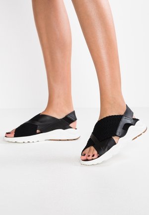 Ben iført sorte sandaler med krydsremme og hvide tykke såler på en hvid gulv mod en ensfarvet baggrund.