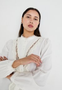 Blouse blanche avec des détails plissés et des accents en dentelle. Présente un col montant, des manches bouffantes et une fermeture à boutons, créant une apparence texturée.