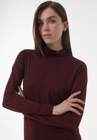 Junge Frau mit glattem braunem Haar, die ein langärmeliges kastanienbraunes Oberteil trägt, die Arme gekreuzt und leicht zur Seite blickend, vor einem weißen Hintergrund.