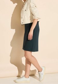 Beige kort denimjacka, mörkgrön knälång klänning med en sidoklyfta och vita sneakers. Slät textur och avslappnad design.